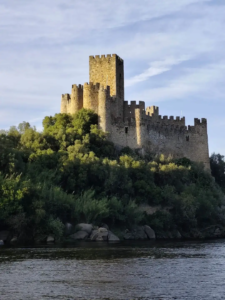 A majestic medieval stone castle perched atop a lush, green rocky islet in the middle of a calm river, under a partly cloudy sky at sunset, with warm golden light illuminating its crenellated towers and walls.