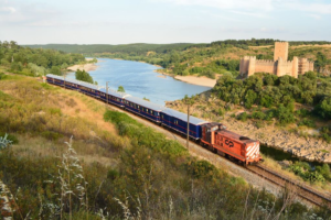A red CP locomotive pulls a long blue luxury passenger train along a scenic railway track beside a wide river, with a historic Almourol Castle perched on a hill in the background under a clear sky.