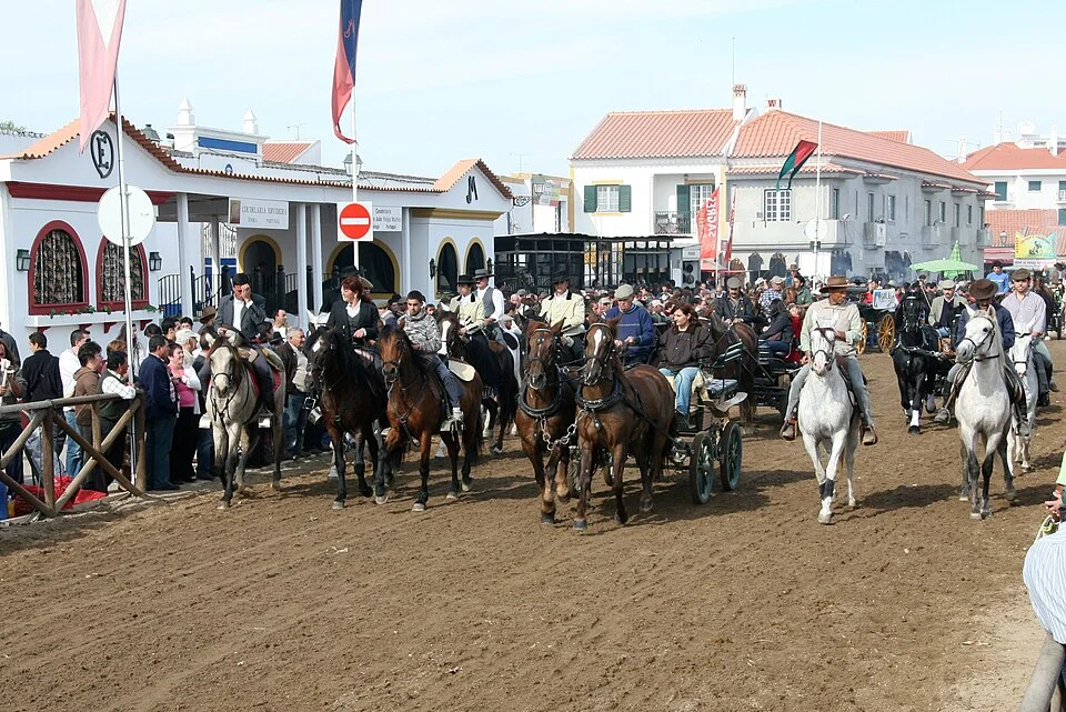 Vista larga do recinto da feira, com cavaleiros, charretes e público junto às casas caiadas da vila.